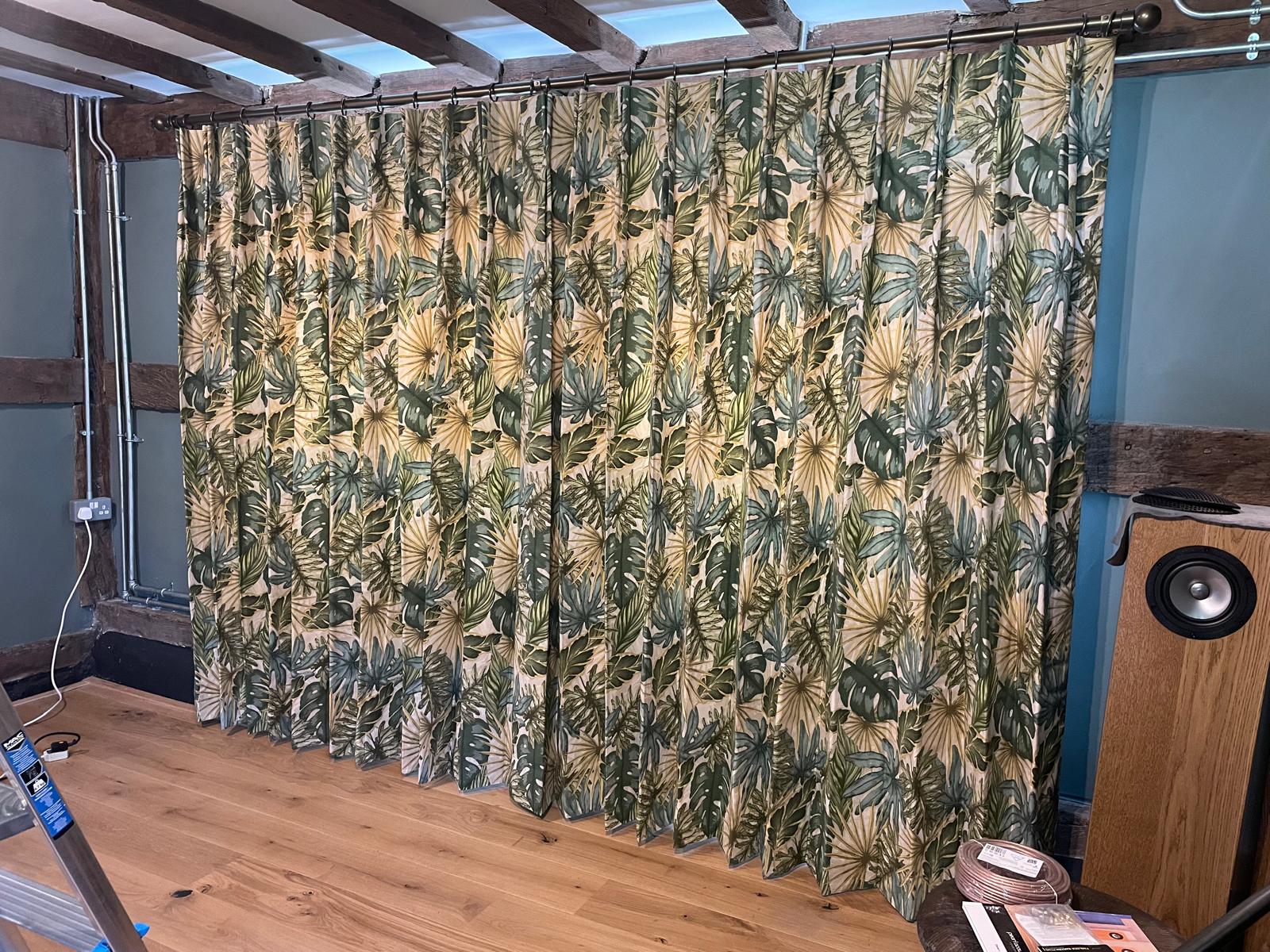 Full-width tropical leaf curtains hung from a brass rod beneath exposed beams