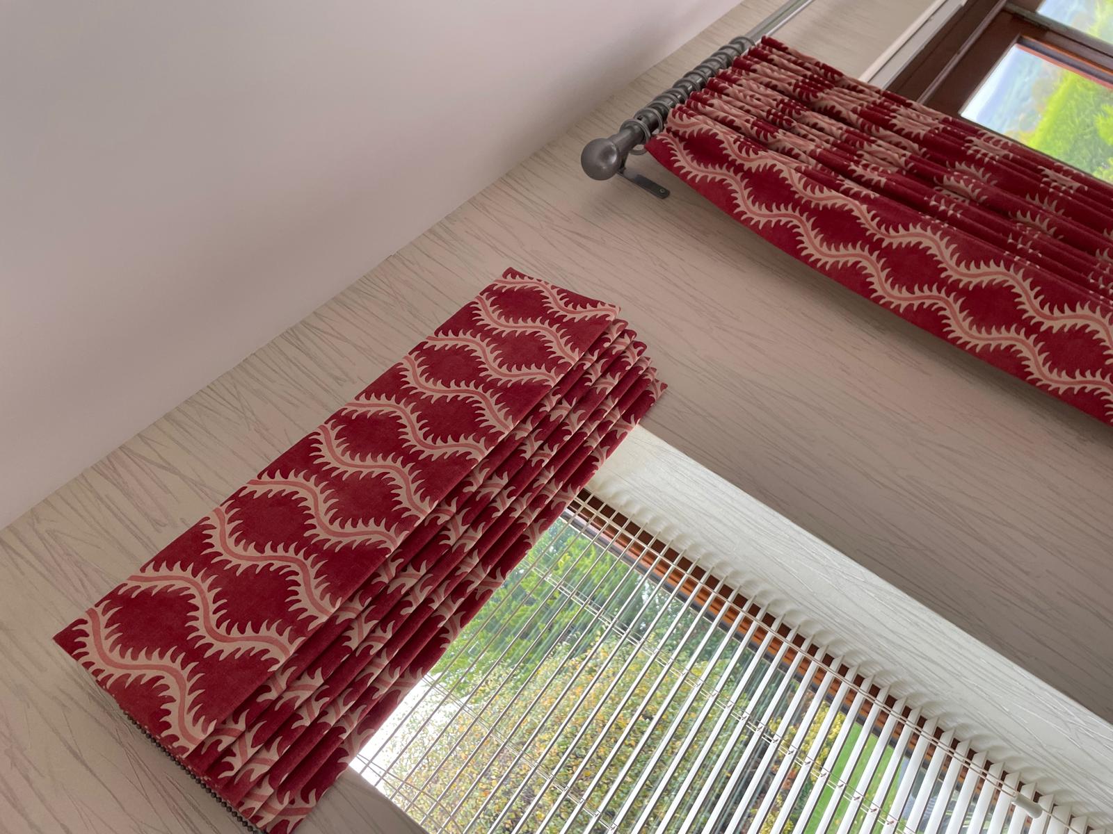 Deep red patterned Roman-style curtain stacked beside a white venetian blind in a bay window
